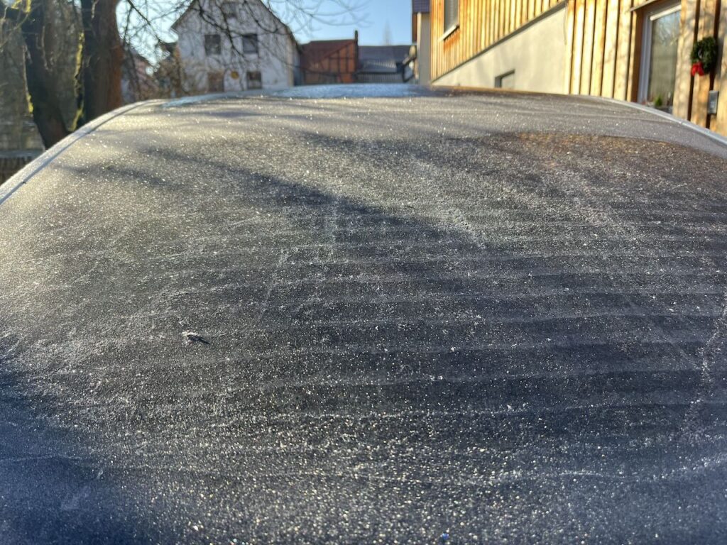 Ein von Raureif überzogene Autoheckscheibe, in Hintergrund ist ein Haus zu sehen. Die Sonne scheint, links steht ein Baum der Schatten auf das Auto wirft.